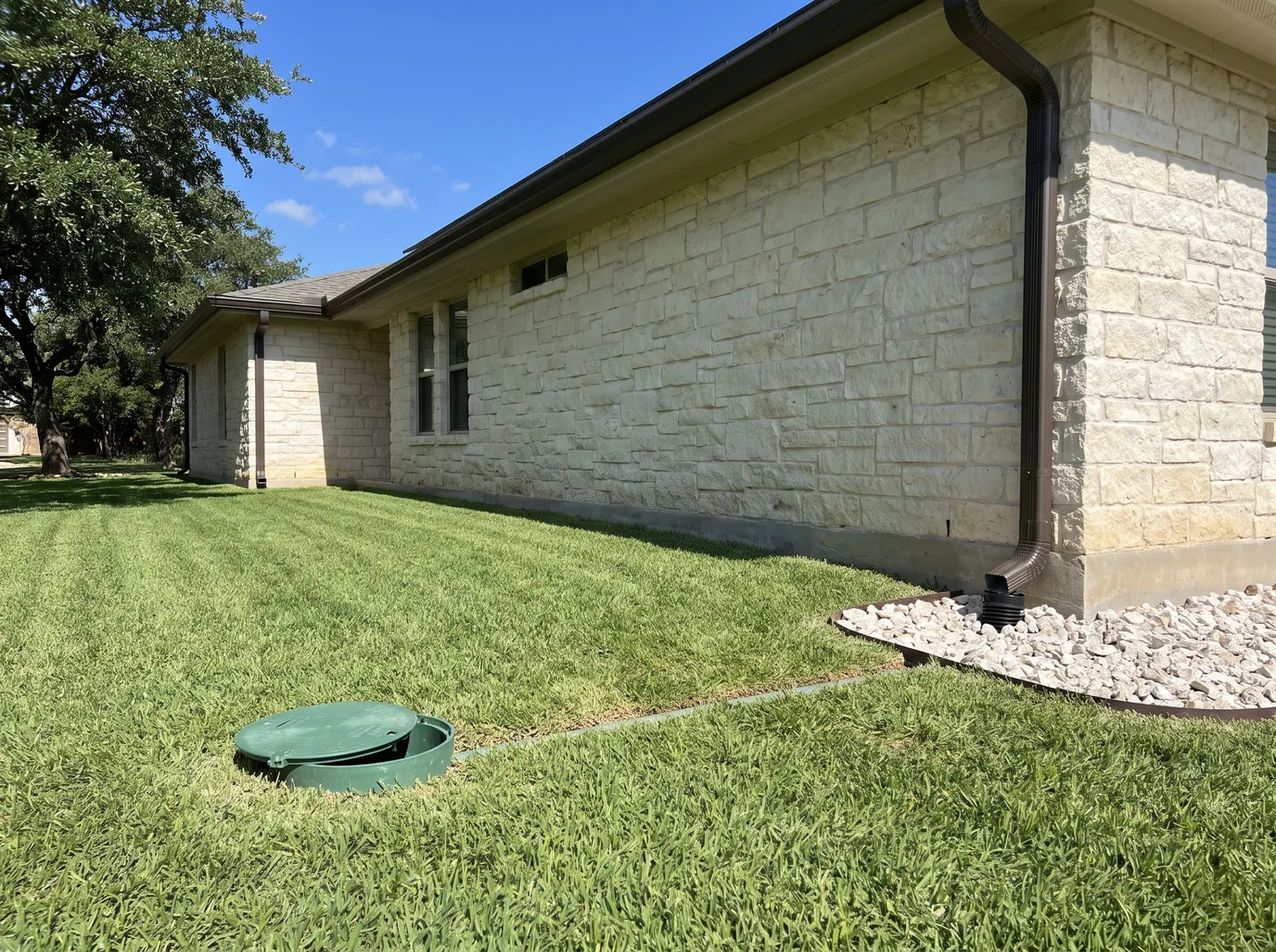 A dark bronze downspout turning into a buried drain line at a limestone house, with a green pop-up emitter out in the lawn