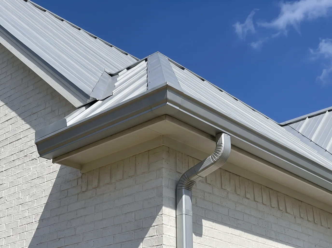 Bare Galvalume steel gutters with a mitered corner and a downspout under a silver standing-seam metal roof