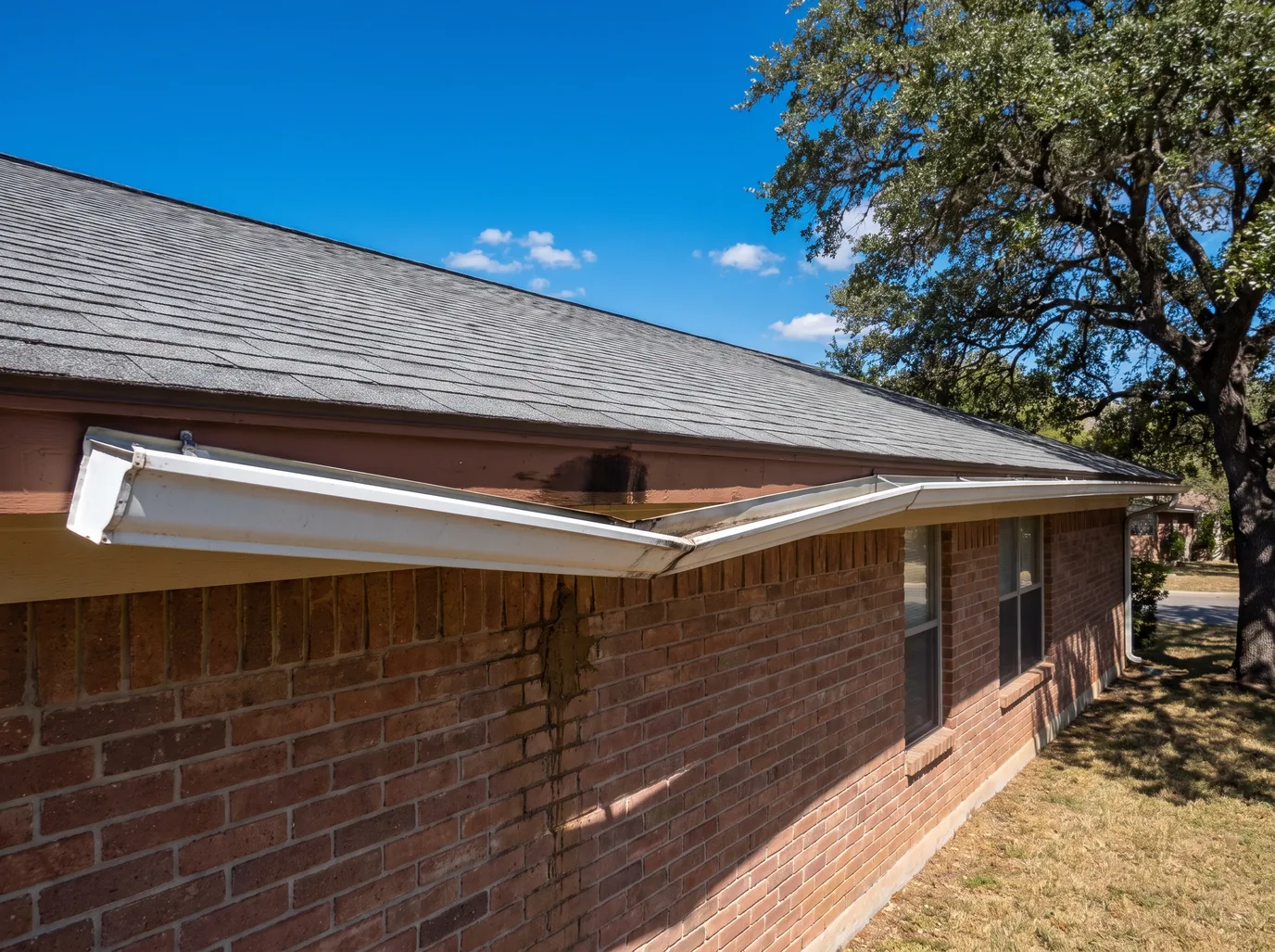An old white sectional gutter sagging away from a water-stained fascia board on a brick ranch house