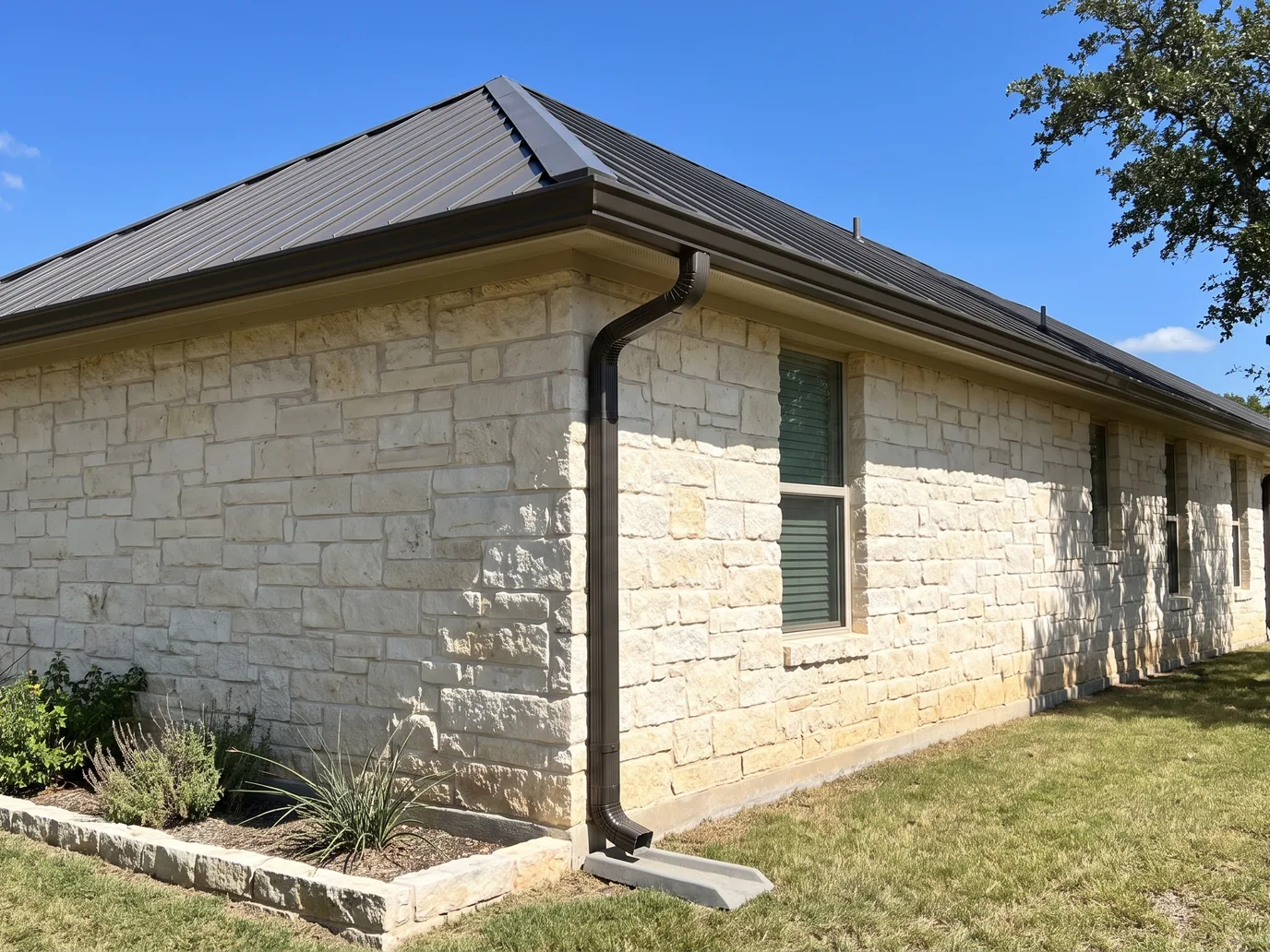 The corner of a limestone house with a dark bronze seamless gutter, a mitered corner, and a downspout that runs down the wall to a splash block