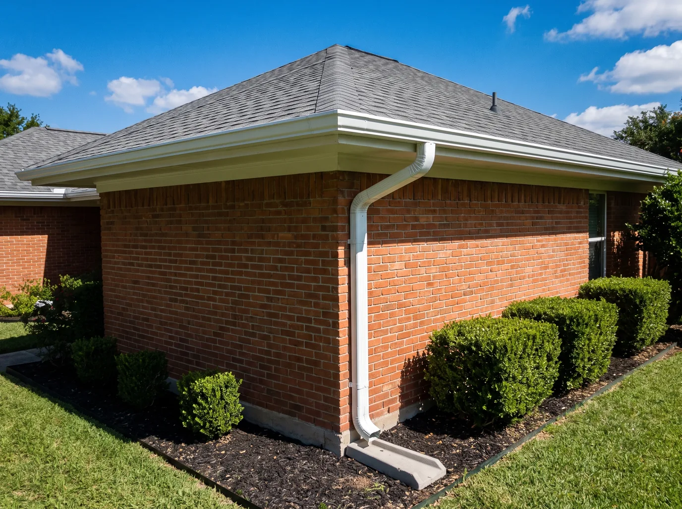 White seamless aluminum gutters and a downspout ending on a concrete splash block at the corner of a red brick ranch house