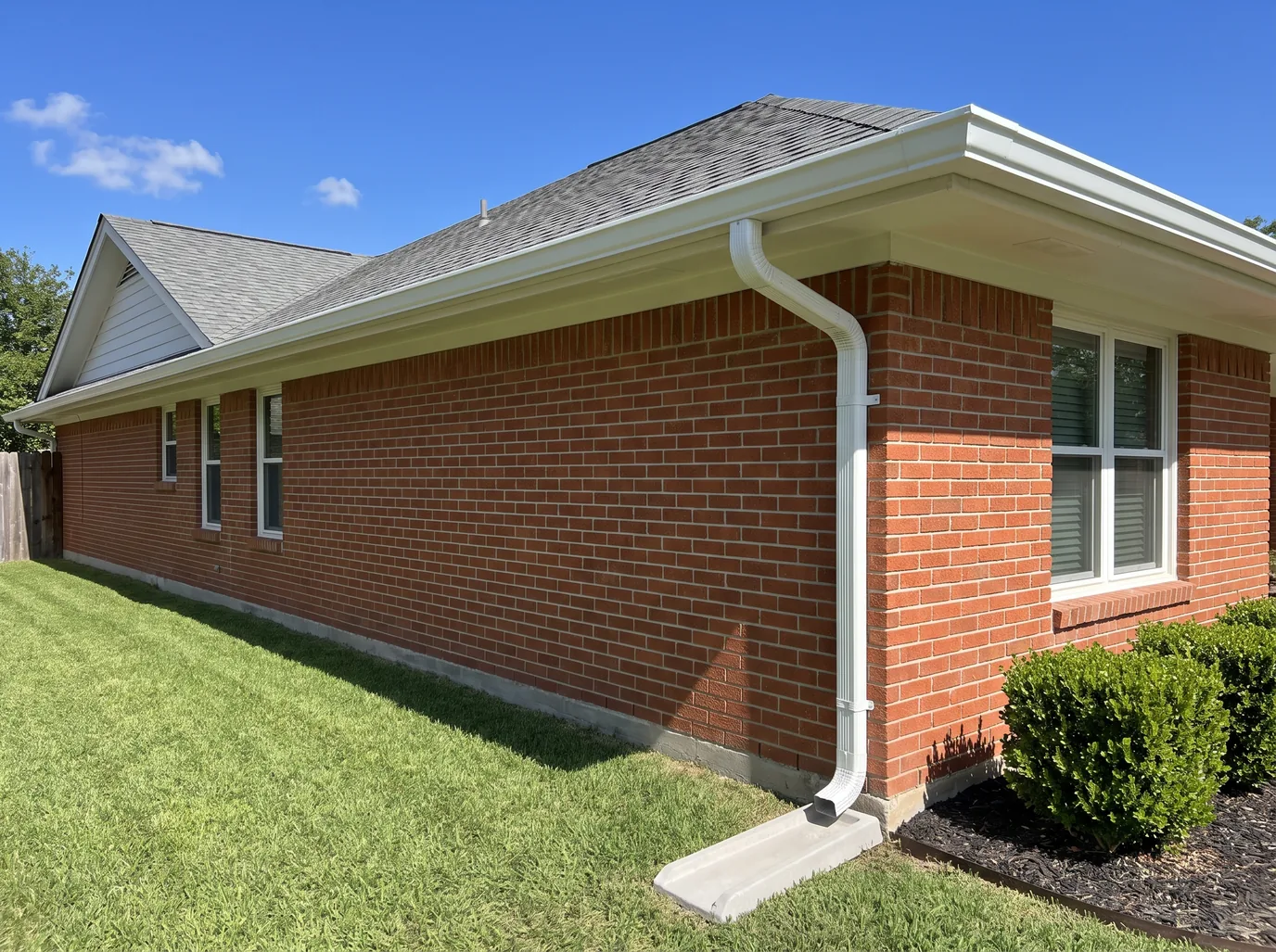 White seamless gutters and a downspout on a sloped splash block at the corner of a red brick ranch house